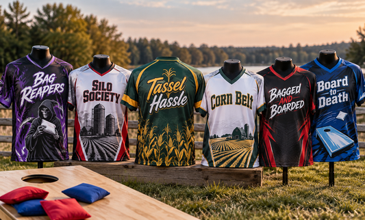 Six custom sublimated cornhole jerseys on mannequins beside boards and bags at dusk